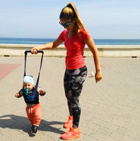 La photo montre une maman sur la plage en train d'apprendre à marcher à son bébé dans son harnais de marche.